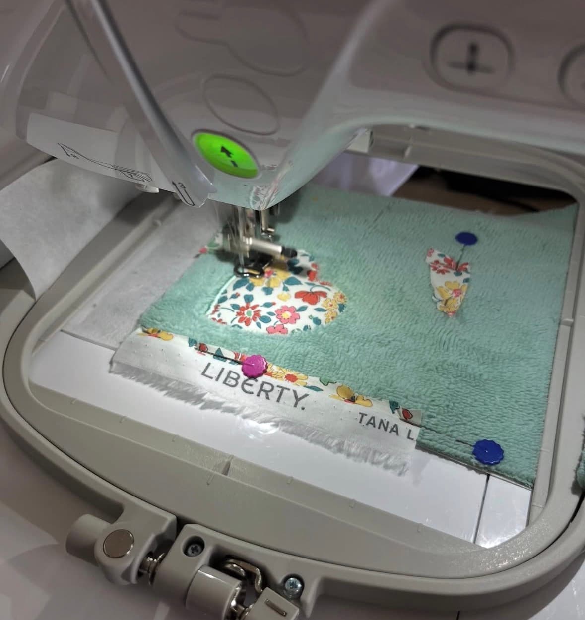 Détail couture : machine à broder posant un appliqué cœur en tissu Liberty sur support vert d’eau.